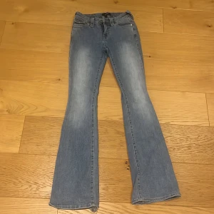 True Religion Becca Bootcut Jeans - Snygga ljusblå bootcut jeans från True Religion, modell Becca Mid Rise Booty. Jeansen har klassiska fickor med lock och knappar bak, samt slitna detaljer vid bensluten. Tillverkade i mjukt jeansmaterial som sitter skönt och ger en avslappnad vibe.