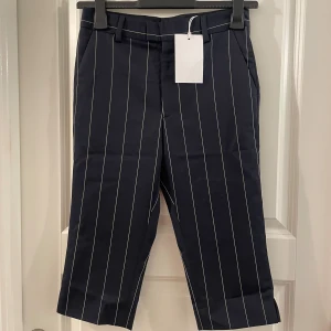 Mörkblå randiga capribyxor - Snygga mörkblå capribyxor med vita vertikala ränder. Byxorna har croppad längd, bälteshällor och sidfickor. Perfekt för dig som gillar en stilren och trendig look. Materialet känns mjukt och har en klassisk känsla.