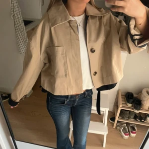 Beige och svart croppad kappa - Snygg croppad kappa i beige och blå med stora fickor framtill och bred krage. Jackan har långa ärmar med blåa detaljer vid muddar och rygg, samt knäppning med två knappar framtill. Perfekt för dig som gillar en loose och trendig stil.