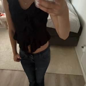 Svart croppad blus med volanger - Säljer en svart croppad blus från GinaTricot med snygga volangdetaljer framtill. Toppen har v-ringad hals och är ärmlös, vilket ger en cool och trendig look. Perfekt för dig som gillar att sticka ut med detaljer och vill ha en topp som känns både modern och stilren. Aldrig använd!