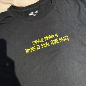 Svart Peanuts t-shirt från Uniqlo - Svart t-shirt från Uniqlo x Peanuts med gul text 'Charlie Brown is trying to steal home base!!' på framsidan och coolt tryck av Peanuts-karaktärer och stjärnor på ryggen. Skön passform och tillverkad i mjuk bomull. Perfekt för dig som gillar serier och streetstyle.