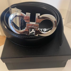 Svart skärp från Salvatore Ferragamo - Snyggt svart bälte från Salvatore Ferragamo med en ikonisk, stor silverspänne i metall. Bältet är tillverkat i slätt skinn och har en stilren, klassisk design som passar till jeans eller kostym. Perfekt accessoar för att lyfta din outfit.