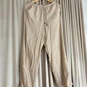 Beiga linnebyxor med snörning - Luftiga beiga linnebyxor med resår i midjan och snörning med trädetaljer. Byxorna har en avslappnad passform och är tillverkade i ett krinklat linnetyg. Perfekta för varma dagar och chill vibes.