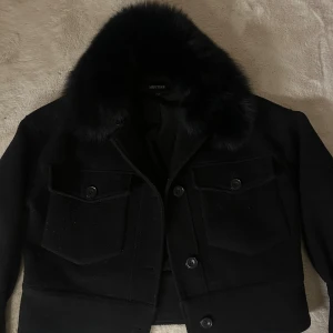 Svart jacka med pälskrage från Meotine - Säljer en svart jacka från Meotine med lyxig pälskrage och stora bröstfickor med knappar. Jackan har en croppad passform och är tillverkad i ullblandning med snygga detaljer. Perfekt för kyliga dagar när du vill vara både varm och stilren.