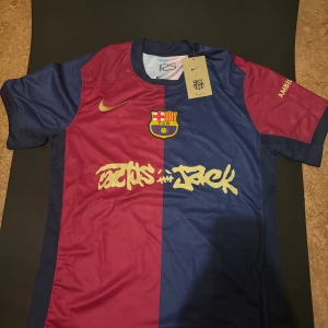FC Barcelona x Nike Travis scott  tröja - Säljer en FC Barcelona fotbollströja från Nike med Cactus Jack-grafik. Tröjan är kortärmad och har klassisk halvdelad design i mörkblått och vinrött, med guldfärgade detaljer och klubbmärke på bröstet. Materialet är lätt och andas, perfekt för match eller träning.