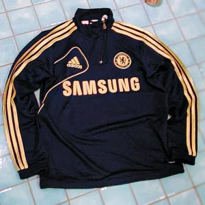 Chelsea FC Track top Adidas fotbollströja [XS/164]  - Chelsea FC långärmad fotbollströja med zip från Adidas i mörkblått med guldiga detaljer och ränder längs ärmarna. Vintage-versionen 2006-2014 i utmärkt skick. Tröjan har halv dragkedja, klubbmärke på bröstet och Samsung som sponsor. Tillverkad i polyester, perfekt för dig som älskar klassisk fotbollsstil.