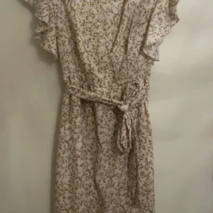 Blommig långklänning  - Långklänning från SHEIN i ljusbeige med små blommiga mönster i gult och brunt. Klänningen har volangärmar, omlottliv och knytskärp i midjan. Perfekt för sommarens alla tillfällen. Använd ett fåtal gånger 