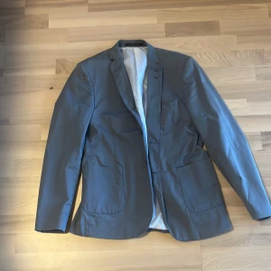 Grå kavaj från Tiger of Sweden - Snygg grå kavaj från Tiger of Sweden med klassisk passform och två framfickor. Kavajen har ljus insida och knappar vid ärmslut. Perfekt för dig som vill ha en stilren och modern look.