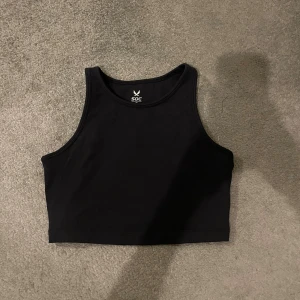 Svart träningslinne - Svart croppad linnetopp från SOC i storlek XS. Toppen har en stilren, sportig look med hög halsringning och cool snörning i ryggen. Materialet är mjukt och stretchigt syntet som andas, perfekt för träning eller chill. Snygg och bekväm att bära till gymmet eller hemma.
