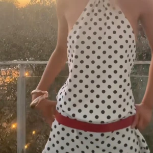 Prickig halterneckklänning - Vit kort halterneckklänning med svarta prickar. Klänningen är ärmlös och har öppen rygg, vilket ger en riktigt snygg och somrig vibe. Perfekt för dig som gillar retrostil och vill sticka ut med ett lekfullt mönster.