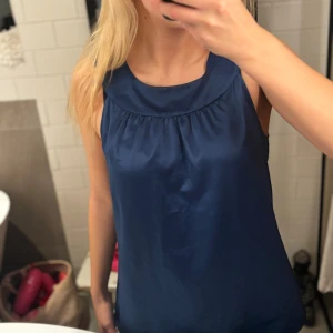 Mörkblå ärmlös linnetopp i satin - Snygg mörkblå linnetopp med rundad hals och lätt glansig satinfinish. Toppen är ärmlös och har ett mjukt fall med diskreta rynkdetaljer vid halsen. Perfekt för dig som gillar stilrena och enkla plagg med lite extra shine.