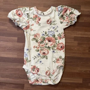 Blommig body - Supersöt body från Newbie i storlek 74 (6-9mån) med puffiga korta ärmar. I extremt god skick. Enda anmärkningen är knäppningen bak på nacken som är smått uttöjd som kan ses på sista bilden, men inget som varken syns eller påverkar funktionen alls!