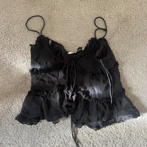 Svart volang linnetopp med snörning - Säljer en svart linnetopp från zara med tunna axelband, volangdetaljer och snörning framtill. Toppen har en lätt transparent, plisserad look och är croppad med vågiga kanter. Perfekt för dig som gillar romantiska och trendiga detaljer.