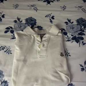 Vit Moncler polotopp med krage - Säljer en klassisk vit polotopp från Moncler i storlek S. Toppen har korta ärmar, krage och två knappar framtill. Moncler-loggan är broderad på bröstet. Materialet är mjuk bomull som känns skönt mot huden. Perfekt för en clean och stilren look.