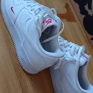 Nike Air Force 1 vita sneakers - Säljer ett par klassiska Nike Air Force 1 sneakers i helvitt skinn med röda detaljer på loggan och häl. Skorna har rund tå, perforerad tåbox och vit platt sula. Perfekta för dig som gillar en clean och tidlös look. Storlek 42 (US 8.5, UK 7.5).