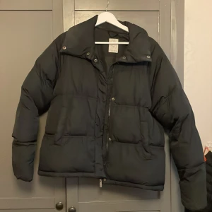 Svart pufferjacka från Etirel - Säljer en svart pufferjacka från Etirel i storlek 36. Jackan har en hög krage, stora fickor och dragkedja framtill. Liten missfärgning på ena sidan av kragen