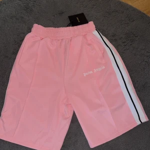 Palm Angels rosa shorts med revär - Snygga rosa shorts från Palm Angels med vit och svart revär längs sidorna. Elastisk midja och sidofickor, samt Palm Angels-logga tryckt på ena benet. Tillverkade i mjuk polyester som känns skön mot huden. Perfekta för en trendig och sportig look.