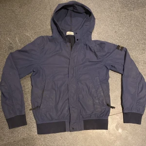 Blå vindjacka Stone Island Junior - Snygg marinblå vindjacka från Stone Island Junior med huva och klassisk patch på ärmen. Jackan har dragkedja framtill, ribbade muddar och två fickor med dragkedja. Perfekt till hösten. Den är två lager (inte fodrad) men det gör att den tål vatten väldigt bra och är varm. Inga defekter men inte tvättad. Såklart äkta då den har certilogo. Den är storlek 154 men jag som är 168 passar den perfekt på som visst på bild så skulle säga XS.