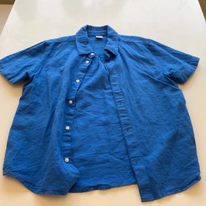 Blå kortärmad skjorta från Lindex - Blå skjorta från Lindex i storlek 146/152, perfekt för dig som gillar en chill och enkel stil. Skjortan har korta ärmar, klassisk krage och vita knappar framtill. Tillverkad i mjukt bomullstyg som känns skönt mot huden.