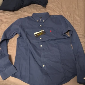 Blå skjorta från Ralph Lauren M - Snygg blå skjorta från Ralph Lauren i klassisk passform. Skjortan har vita knappar, lång ärm och den ikoniska röda Polo-loggan broderad på bröstet. Passar mig som är ungefär 180 väldigt bra men råkade beställa 2 när jag skulle beställa en så fick en över 