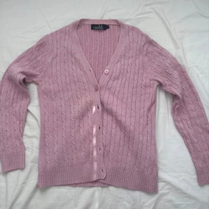 Ljusrosa kabelstickad kofta Isolde - Supersöt ljusrosa kabelstickad kofta från Isolde med v-ringning och knappar framtill. Koftan har långa ärmar, ribbade muddar och är gjord i mjukt stickat material. Perfekt att slänga över en topp för en chill look.  Koftan har ett hål på baksidan runt kragen, inget som syns och kan sys ihop.   Sista bilden är för att visa att alla knappar finns där, färgen ser annorlunda ut men det är de första bilderna som visar riktiga färgen