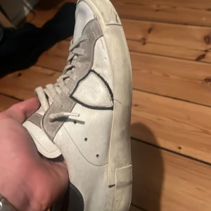 Vita och grå sneakers i skinn - Säljer ett par vita sneakers med grå detaljer och snörning. Skorna har en klassisk låg profil, rund tå och platt sula. Ovandelen är i skinn med kontrasterande grå paneler och svart kant på hälen. Perfekta för dig som gillar en stilren och enkel look. Är lite slitna då man inte ser nånting. Passar 38 
