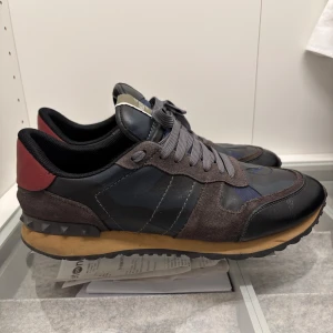 Valentino Garavani Rockrunner sneakers - Snygga Valentino Garavani Rockrunner sneakers i mörkgrått, svart och marinblått med röda detaljer på hälen. Skorna har camouflagemönster, snörning och coola nitar på sulan. Box medföljer!