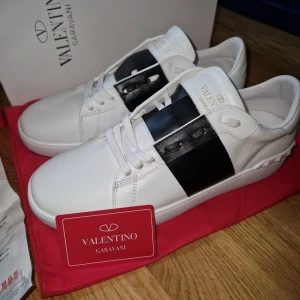 Valentino Garavani Open sneakers vit/svart - Snygga Valentino Garavani Open sneakers i vitt skinn med bred svart läderdetalj över vristen. Klassisk rund tå, vita snören och diskret logga i guld på plösen. Perfekta för dig som gillar stilrena och lyxiga sneakers med en unik twist.