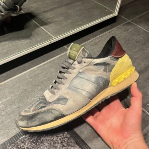 Valentino Garavani Rockrunner sneakers - Säljer ett par Valentino Garavani Rockrunner sneakers med grått och blått camouflage-mönster, detaljer i gult och vinrött på hälen samt grönt märke på plösen. Skorna har snörning, platt sula och är tillverkade i mocka och skinn. Perfekt för dig som gillar streetstyle med lyxig touch.