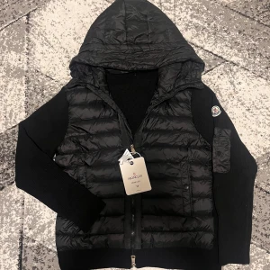 Svart Moncler pufferjacka med huva - Svart pufferjacka från Moncler med stickade ärmar och huva. Jackan har dragkedja framtill, Moncler-logga på ärmen och coolt serietryck på insidan. Materialmix med dunfyllning och stickad polyester. Perfekt för dig som vill ha en snygg och bekväm jacka med streetkänsla. NFC funkar även när man scanna loggan 