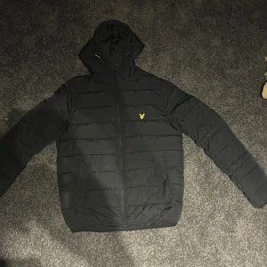 Svart pufferjacka från Lyle & Scott - Typ aldrig använt. Svart pufferjacka från Lyle & Scott med huva och gul logga på bröstet. Jackan har quiltad design, dragkedja framtill och långa ärmar. Perfekt för kalla dagar och har en clean, stilren look som funkar till streetwear.