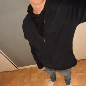 Mörkblå overshirt från Dressmann M - Mörkblå overshirt från Dressmann i storlek M. Jackan har klassisk krage, två bröstfickor med knappar och dragkedja framtill. Materialet är mjukt och känns som bomull, perfekt för lager-på-lager. Enkel och stilren design som funkar till jeans.