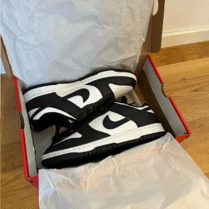 Nike Dunk Low svart/vit sneakers - Säljer ett par Nike Dunk Low sneakers i klassisk svart och vit färg. Skorna har en låg siluett, svart snörning och vit sula. Tillverkade i skinn med perforerad tåbox för extra ventilation. Ikonisk Swoosh-logga på sidorna och Nike-brodyr på hälen. Perfekta för en clean streetstyle.