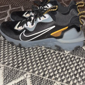 Nike React vision  Element 55 sneakers svart/grå - Nike React Element 55 sneakers i svart med grå och gula detaljer. Skorna har en chunky grå sula, reflekterande partier och klassisk vit Swoosh på sidan. Ovandelen är i syntetmaterial med svarta snören och coola mönstrade dragflikar. Perfekta för dig som gillar streetstyle. I princip nyskick och knappt använda.