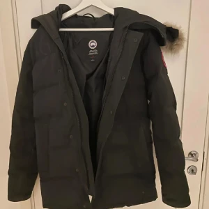 Svart dunjacka från Canada Goose - Säljer en svart dunjacka från Canada Goose med huva och snygg pälskant. Jackan har klassisk patch på ärmen, dragkedja och knappar framtill. Perfekt för kalla dagar och riktigt varm. Stilsäker och tidlös modell med lång ärm.