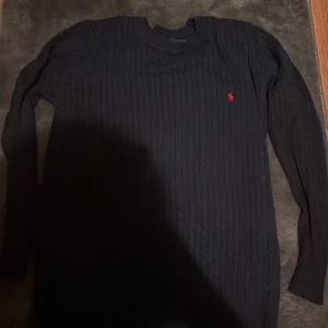 Mörkblå stickad tröja Polo Ralph Lauren - Mörkblå stickad tröja från Polo Ralph Lauren med klassiskt röd broderad logga på bröstet. Tröjan har rund halsringning, ribbade muddar och ett stilrent, ribbat mönster. Perfekt för dig som gillar en clean och tidlös look. 