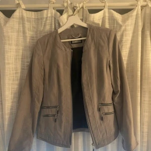 Beige mockajacka med dragkedjor - Snygg beige mockajacka från Vero Moda med stilrena detaljer och rund hals. Jackan har två par dragkedjefickor framtill och är helfodrad. Perfekt för dig som gillar en clean look med lite edge.