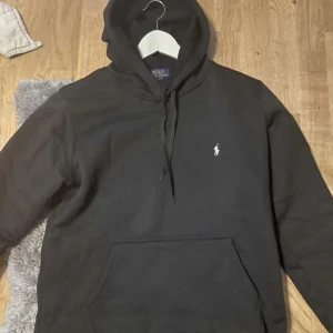 Svart hoodie från Polo Ralph Lauren - Svart hoodie från Polo Ralph Lauren med klassisk vit logobrodyr på bröstet. Tröjan har huva med snörning och en stor magficka framtill. Perfekt för dig som gillar stilren och enkel design med premiumkänsla.
