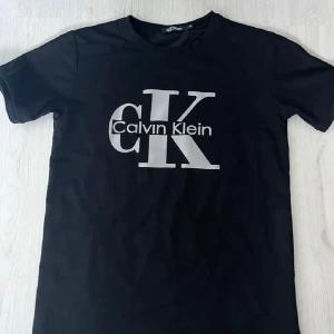 Svart Calvin Klein t-shirt med tryck - Snygg svart t-shirt från Calvin Klein med stort CK-logotryck i silver på bröstet. Klassisk rund hals och korta ärmar, perfekt för en clean och modern look. Materialet känns mjukt och skönt mot huden.