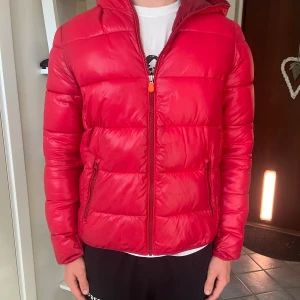 Röd pufferjacka från Save The Duck - Säljer en röd pufferjacka från Save The Duck med huva och dragkedja. Jackan är tillverkad i vattenavvisande syntetmaterial med Plumtech-fyllning, helt animal free. Har Save The Duck-logga på ärmen och två sidofickor med dragkedja. Perfekt för kalla dagar. Storleken är 16 years vilket motsvarar S.