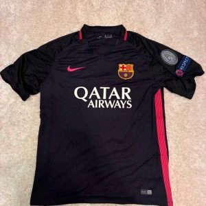 FC Barcelona Neymar Jr 11 matchtröja - Säljer en FC Barcelona matchtröja från Nike med Neymar Jr och nummer 11 på ryggen. Tröjan är svart med rosa detaljer, klubbmärke på bröstet, Qatar Airways tryck och Champions League-patch på ärmen. Tillverkad i lätt, ventilerande polyester med korta ärmar.