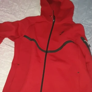 Röd Nike vindjacka med huva - Nike vindjacka i rött med svarta detaljer och hel dragkedja framtill. Jackan har huva, svarta linjer över bröstet och ärmen samt en ficka med dragkedja på armen. Perfekt för dig som gillar sportig stil och vill sticka ut.