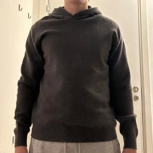 Mörkgrå merinoull tröja från vailent - Mysig mörkgrå stickad hoodie med huva och långa ärmar. Tröjan har en enkel design med ribbade muddar vid ärmslut och nederkant. Perfekt för dig som gillar en chill och stilren look. Passar dig som vill ha något bekvämt och snyggt till hösten eller vintern.