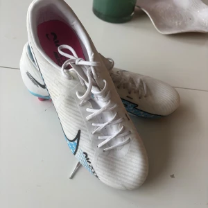 Nike Air Zoom Vapor vita fotbollsskor - Nike Air Zoom fotbollsskor i vitt med blå och svarta detaljer samt rosa och svarta dobbar. Snyggt mönstrad sula och stor Nike-logga på sidan. Skorna har snörning och är designade för grymt grepp på planen. 