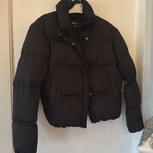 Svart pufferjacka från FB Sister XS - Svart pufferjacka från FB Sister i storlek XS. Jackan har hög krage, två fickor med knappstängning och dragkedja framtill. Den är quiltad och har en kortare, boxig passform som ger en cool vibe. Perfekt för kalla dagar när du vill vara både varm och snygg.