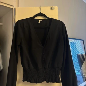 Svart blus med knappar och smock - Svart blus från nelly med v-ringning och klädda knappar framtill. Smockad och rynkad nederkant samt ärmslut ger en snygg och trendig siluett. Passar perfekt  för dig som gillar stilrena och coola plagg för en utekväll