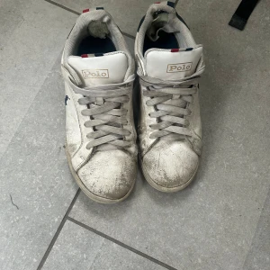 Vita sneakers från Polo Ralph Lauren - Säljer ett par vita sneakers från Polo Ralph Lauren med grå snörning och rund tå. Skorna har Polo-logga på sidan och sula, samt diskreta blå och röda detaljer på plösen. Tillverkade i skinn med platt sula och klassisk sneakerform.