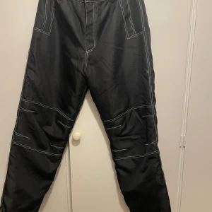 Svarta bluewear byxor med foder o kontrastsömmar - Svarta bluewear fodrade byxor med vita kontrastsömmar och stora fickor både fram och bak. Byxorna har en loose passform och är tillverkade i ett slitstarkt syntetmaterial. Snygga detaljer med extra sömmar längs benen och praktiska fickor för en cool och funktionell look. Fodrade och bra på vintern. Både för herr o dam. 