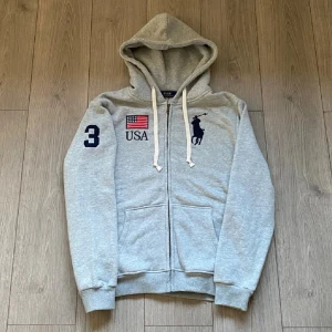 Grå hoodie från Polo Ralph Lauren - Snygg grå hoodie från Polo Ralph Lauren med dragkedja, broderad USA-flagga och stor logga på bröstet. Nummer 3 på ärmen och vita snören i huvan. Perfekt för dig som gillar sportig och klassisk stil (betecknad som storlek L men passar M)