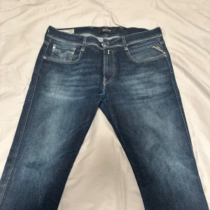 Replay Anbass jeans blå strl 34/32 - Snygga Replay Anbass jeans i mörkblå tvätt med klassisk femficksdesign och kontrastsömmar. Modellen har rak passform och normal midja. Materialet är slitstarkt jeans med en mjuk känsla. Perfekt för dig som gillar stilrena och tidlösa jeans.